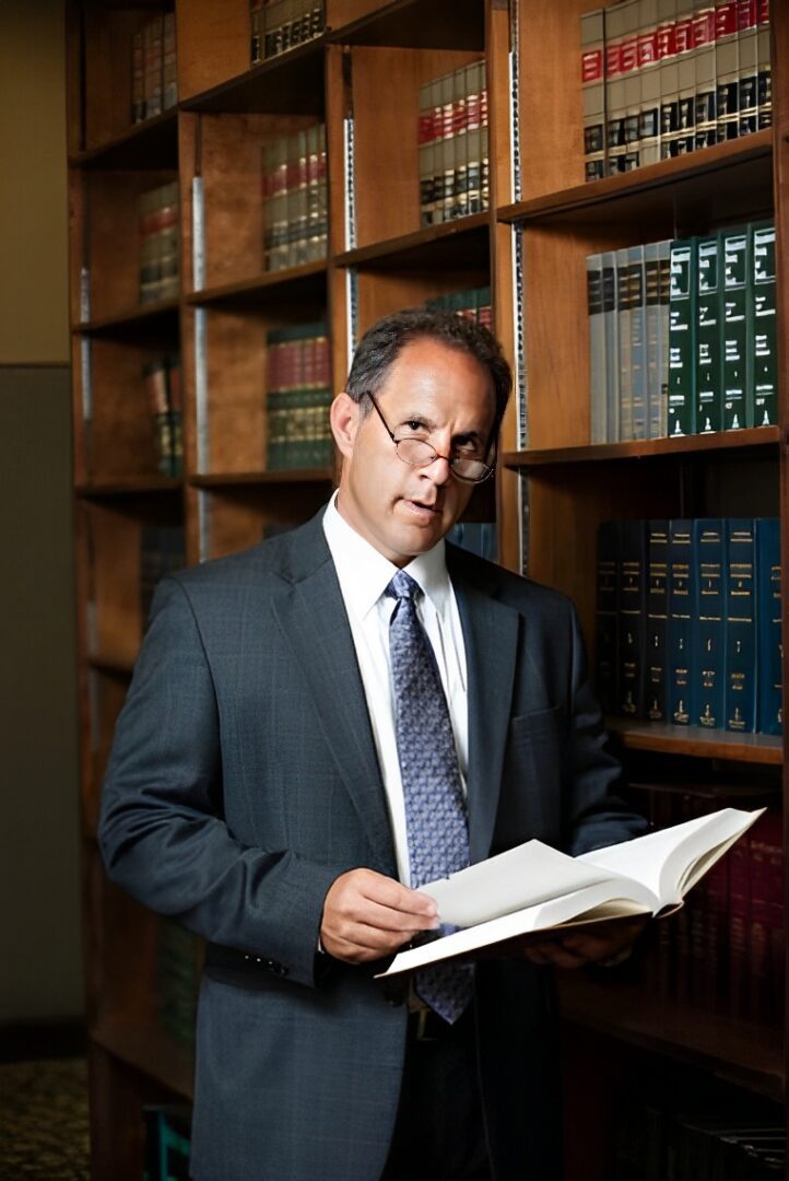 Man in a suit reading a book in a library, standing in front of shelves filled with books.