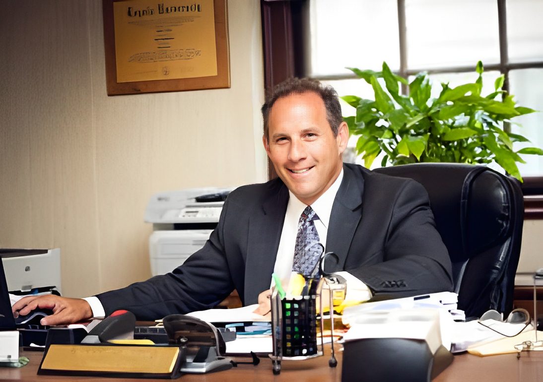 A man in a suit sits at a cluttered desk in an office, smiling at the camera.