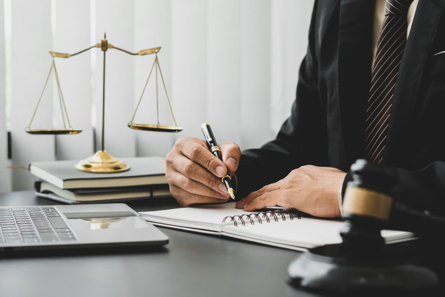 A person in a suit writes in a notebook at a desk with scales of justice, a gavel, and a laptop.