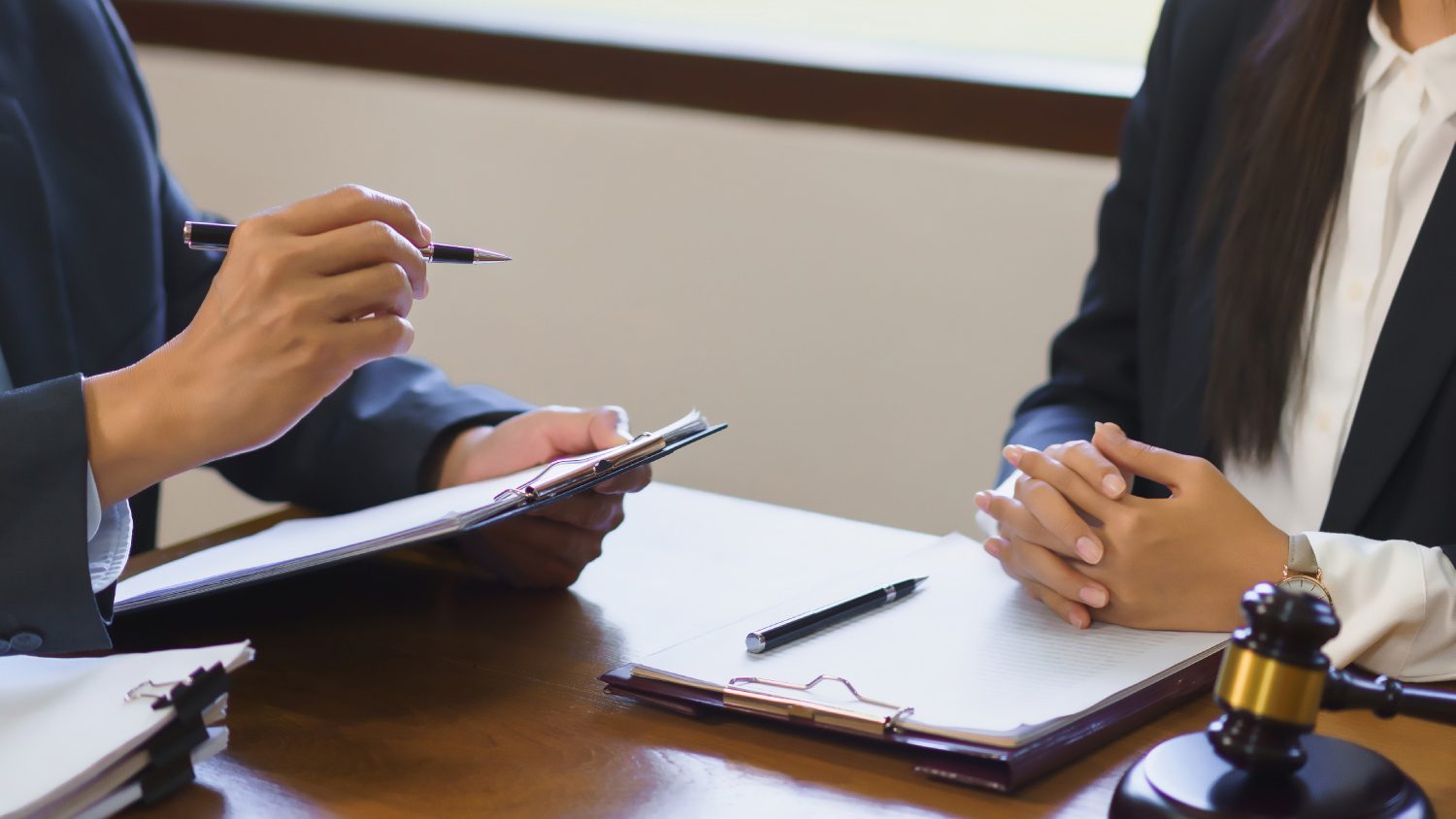 Two people in business attire are sitting at a table with documents, a pen, and a gavel. One holds a clipboard and pen, while the other has their hands clasped on the table.