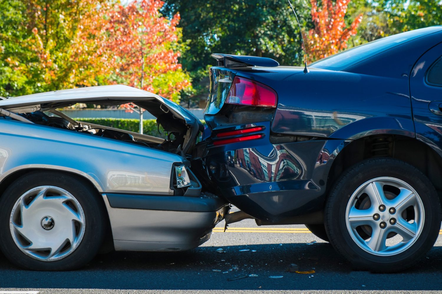 Two cars involved in a rear-end collision on a road. The front of a silver car is damaged against the back of a black car, with fall foliage visible in the background.