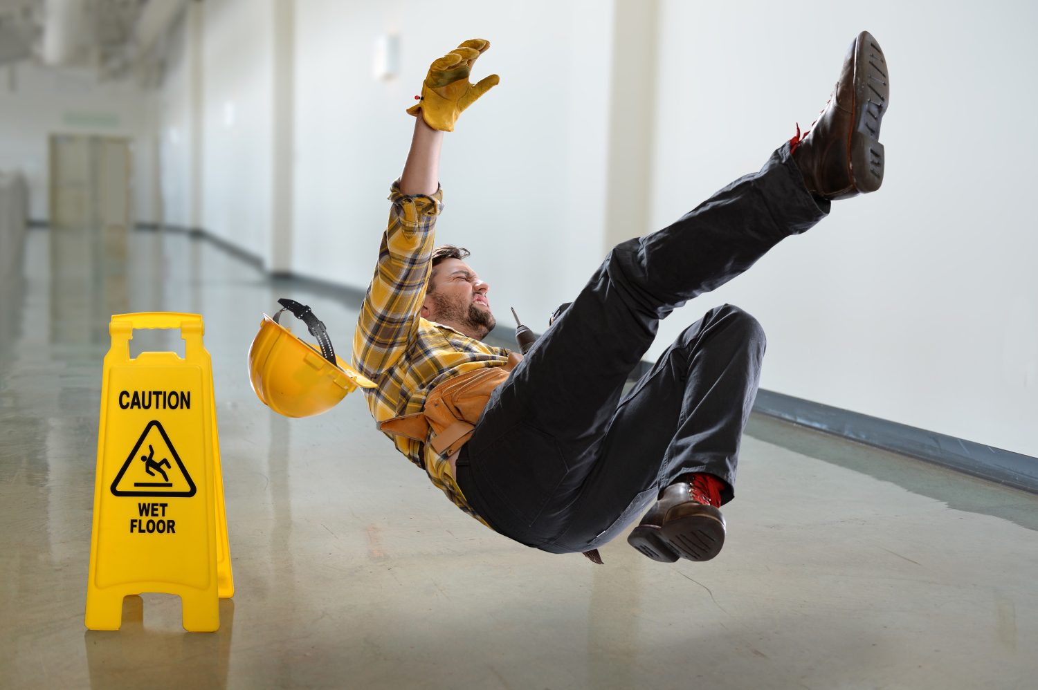 Worker slipping on wet floor beside caution sign, wearing a plaid shirt, gloves, and safety helmet.