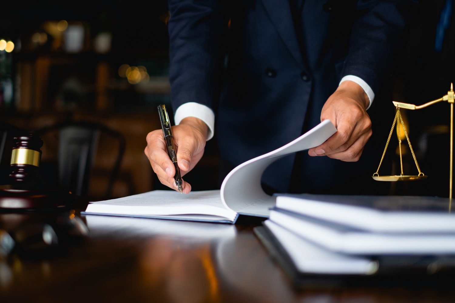 A person in a suit signs a document at a desk with a gavel, scales of justice, and stacked books nearby.